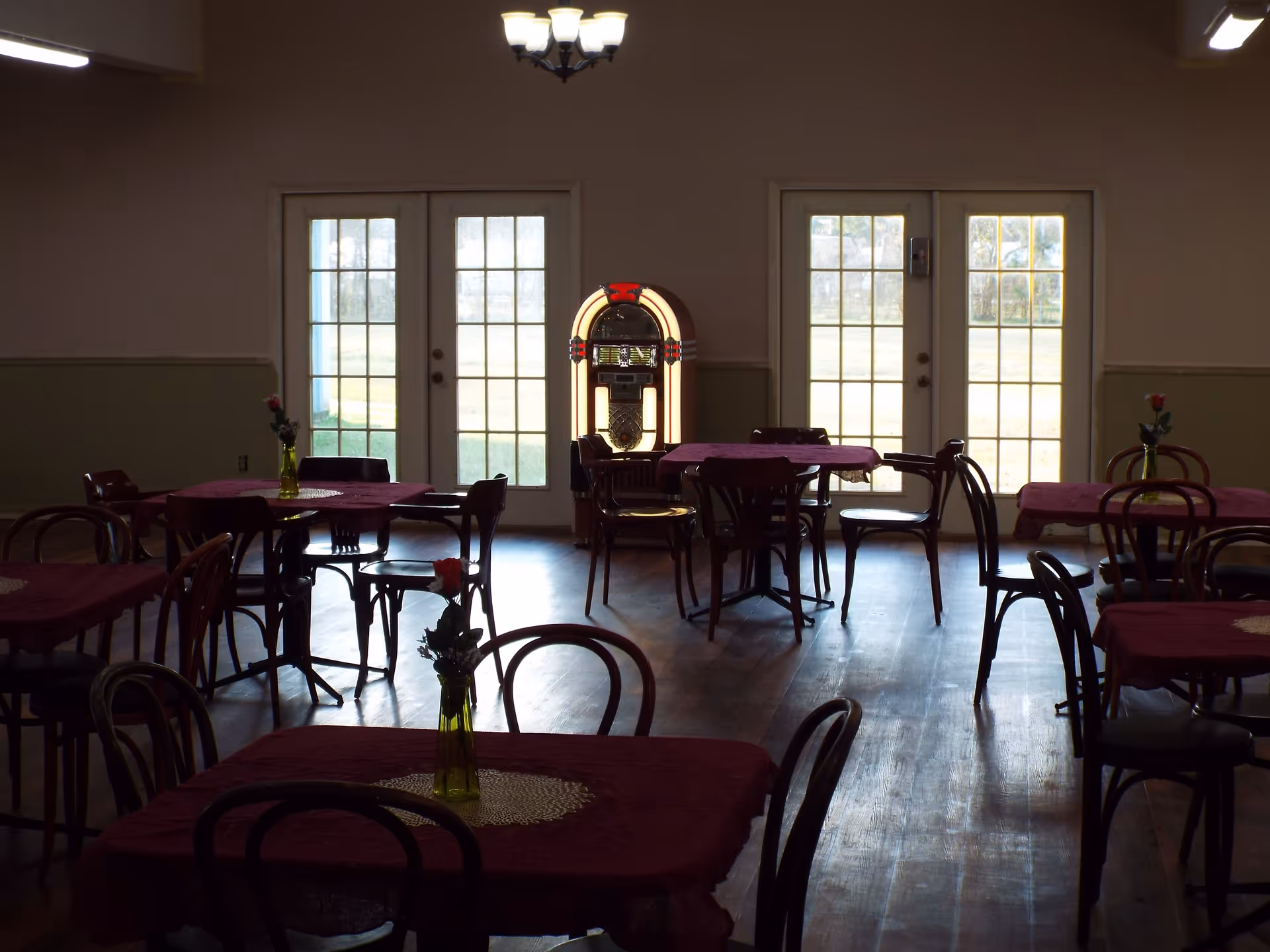 Interior view of a dining room with several tables covered with red tablecloths and each table has a small vase with a single red flower. There are wooden chairs around the tables and a jukebox is positioned against the wall between two sets of French doors letting in natural light.