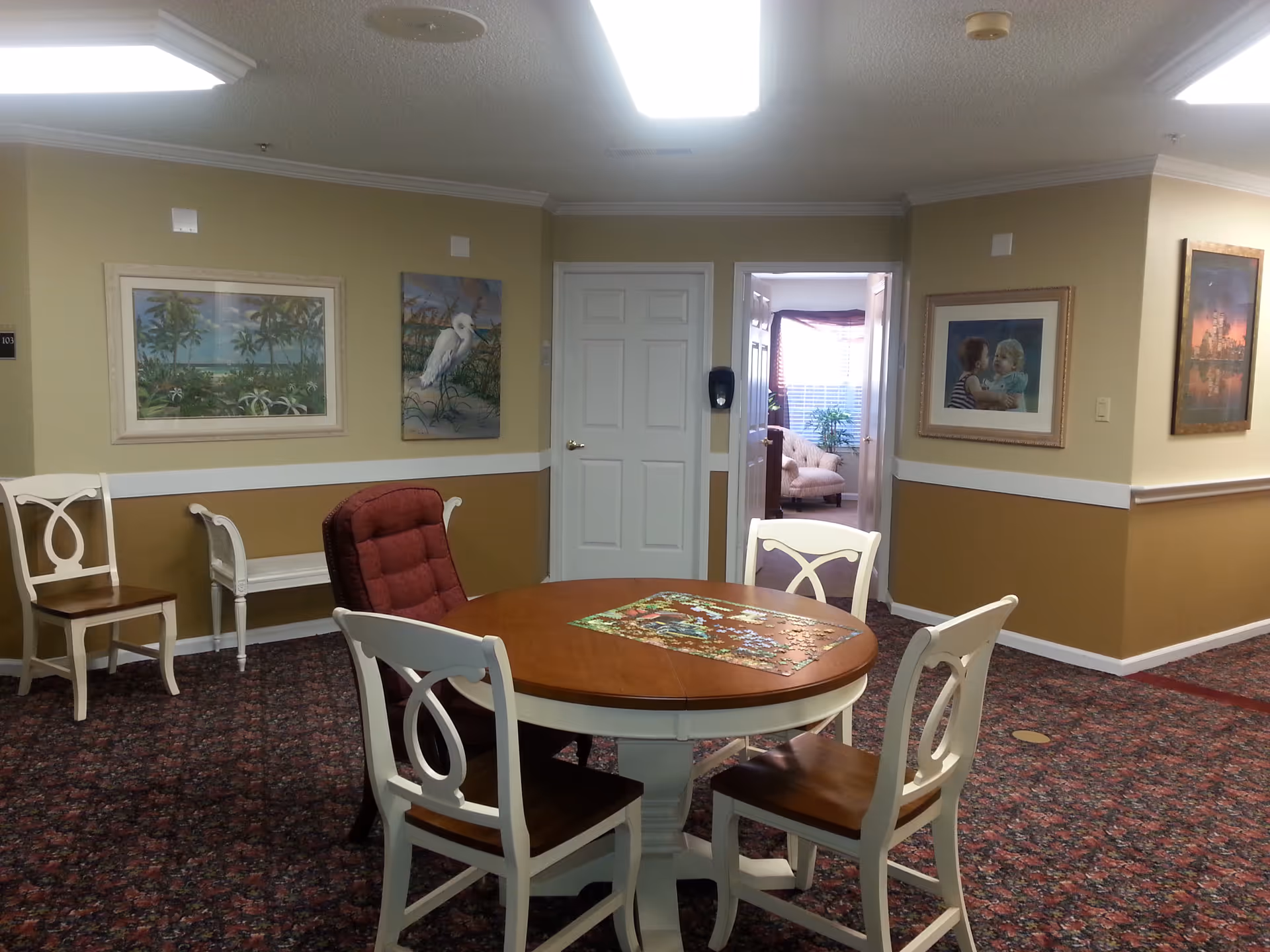 Round wooden table with four chairs and a partially completed puzzle in a carpeted common room with framed artwork and a doorway to a sitting area.