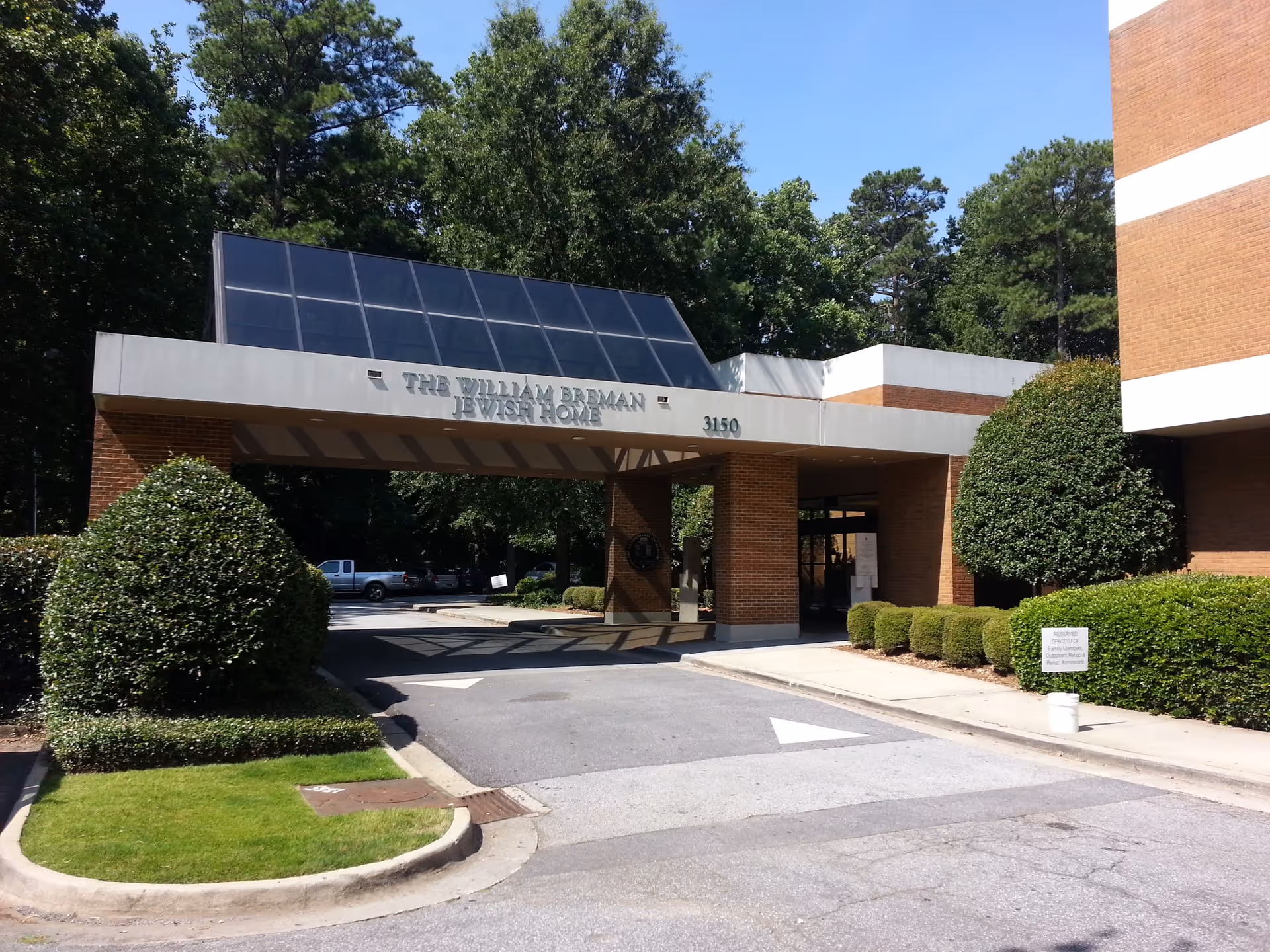 Entrance to The William Breman Jewish Home building with a covered driveway, surrounded by neatly trimmed bushes and trees under a clear blue sky.
