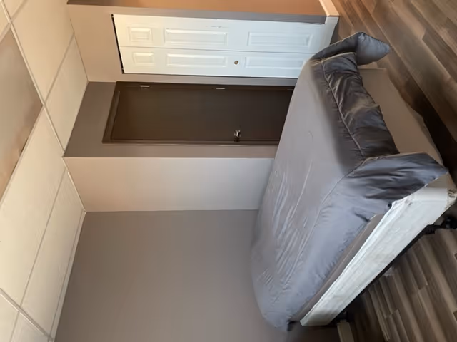 A simple bedroom with a bed covered in a gray comforter, a white closet door, and a brown entry door. The room has gray walls and wood-patterned flooring.