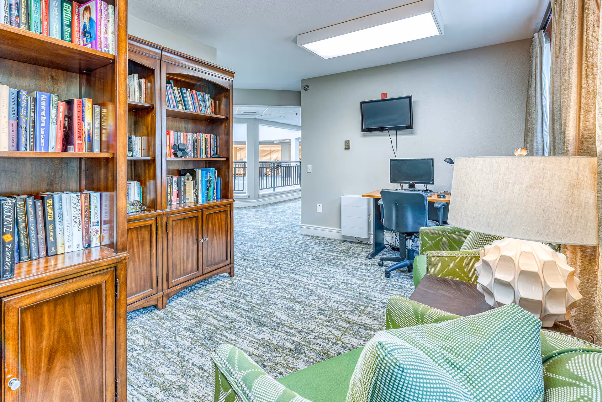 Interior view of a senior living facility common area featuring wooden bookshelves filled with books on the left, a workspace with a computer and two monitors on the right, and green patterned armchairs with a decorative lamp in the foreground. The area has carpeted flooring and a large window with curtains allowing natural light.