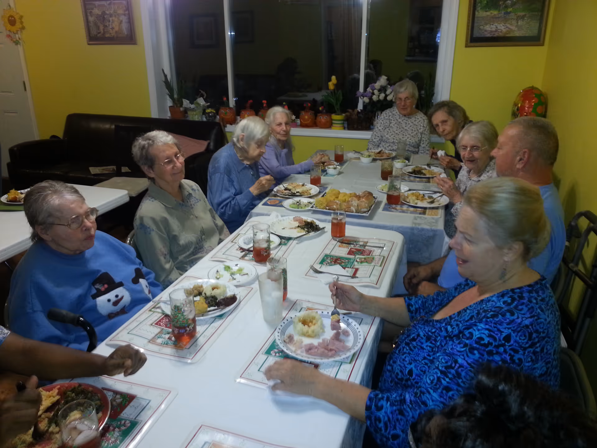 A group of elderly people sitting around a long dining table enjoying a meal together in a brightly lit room with yellow walls. The table is set with plates of food, glasses of drinks, and festive placemats. There are decorations on the windowsill behind them, including small pumpkin figures and potted plants.