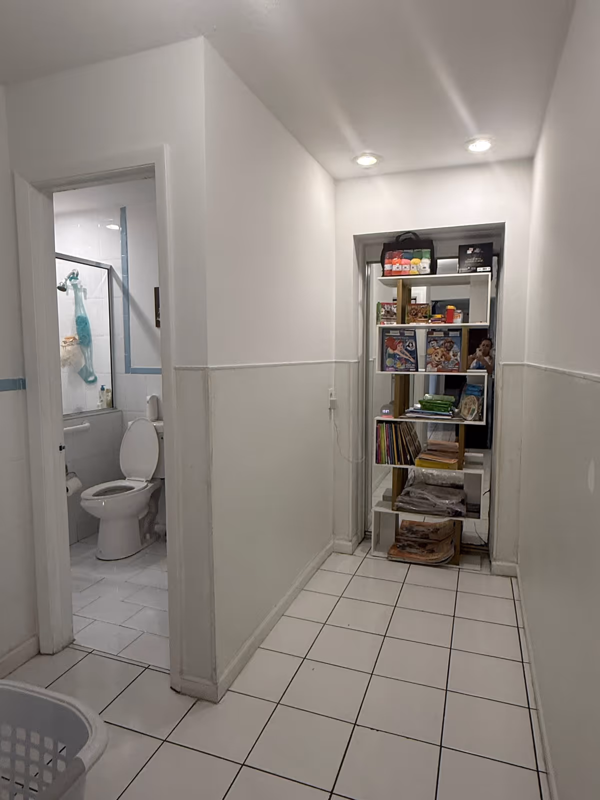 A narrow hallway with white tiled floor and white walls. On the left side, there is an open door leading to a bathroom with a toilet and a shower area visible. At the end of the hallway, there is a shelving unit filled with various items including books, boxes, and other small objects. Two ceiling lights illuminate the hallway.
