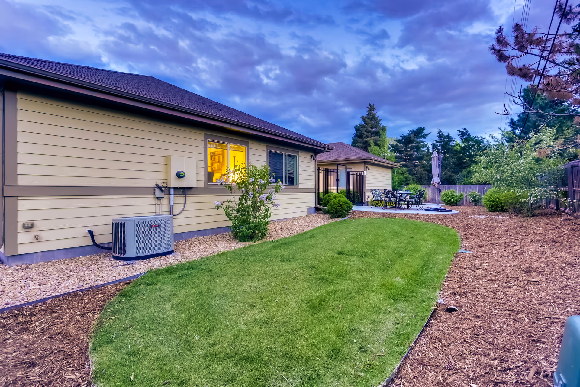 Outdoor view of a single-story building with beige siding and a dark roof, featuring a green lawn, bushes, and a patio area with outdoor furniture and an umbrella under a cloudy sky.