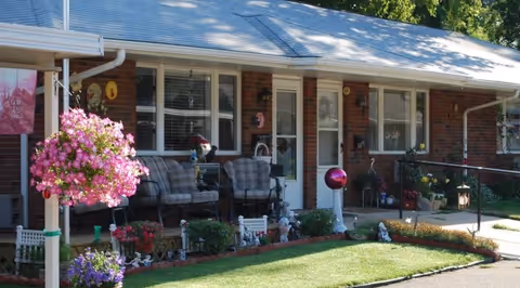 Front porch of a single-story brick senior living home with chairs, hanging flower baskets, potted plants, and a small landscaped lawn.