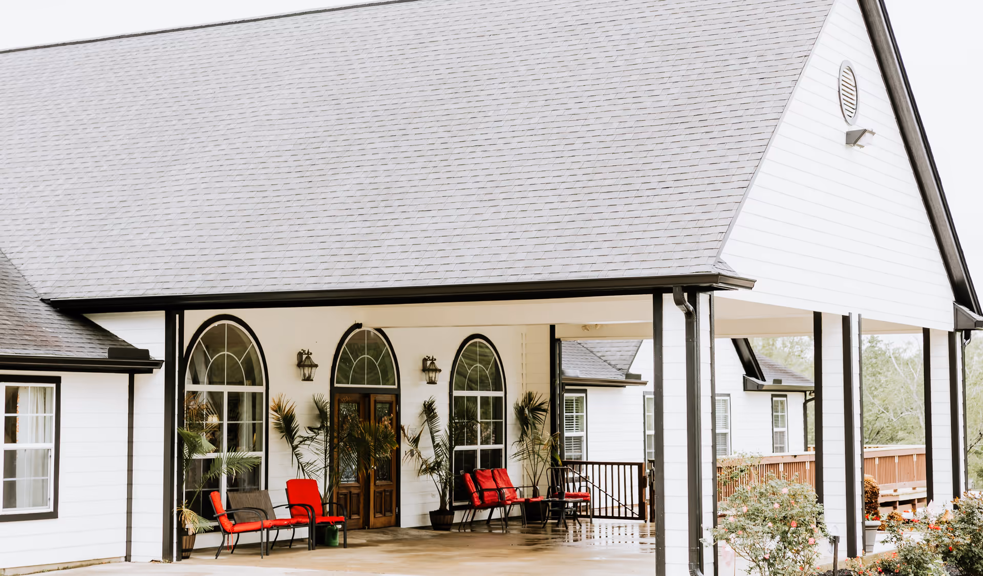 Covered front porch of a white building with arched windows, potted plants and red chairs.