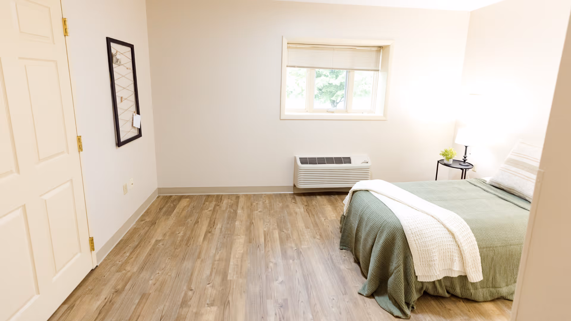 A simple, clean bedroom with light-colored walls and wood flooring. The room features a bed with green bedding and a white throw blanket, a small round side table with a lamp and a small plant, a window with blinds, and a wall-mounted air conditioning unit below the window. A framed mirror hangs on the wall near a closed door.