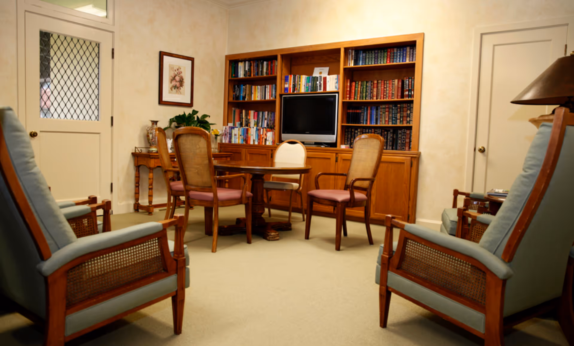 A comfortable common room with upholstered armchairs arranged around a small table, a bookcase with a TV, and wooden furniture.