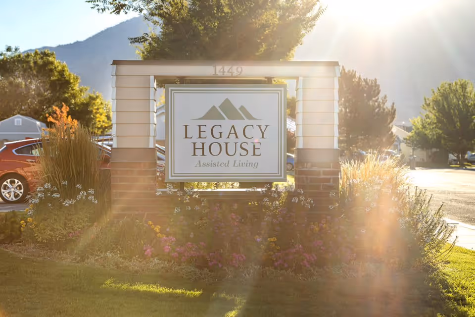 A sign for Legacy House Assisted Living mounted on a brick and white wooden frame structure, surrounded by flowers and greenery, with sunlight creating a lens flare effect. Trees, parked cars, and a mountain are visible in the background.