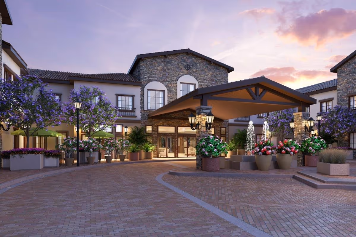 Exterior view of Varenita Westlake facility at sunset, featuring a stone and stucco building with a covered entrance, decorative street lamps, potted plants, flowering trees, and water fountains in the courtyard.