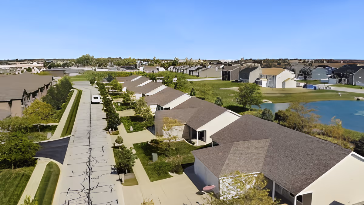 Aerial view of a residential community with single-story homes arranged along a curved road. The area features green lawns, trees, and a small pond on the right side under a clear blue sky.
