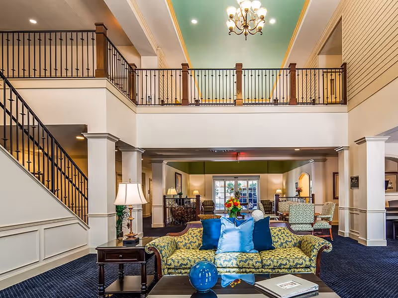 A spacious and elegant living room area in The Windsor at Ortega featuring a patterned sofa with blue pillows, a coffee table with decorative items, a side table with a lamp, and a staircase with black railings leading to an upper balcony. The room has high ceilings with a chandelier and a light blue ceiling accent, with additional seating and lamps in the background near large windows.