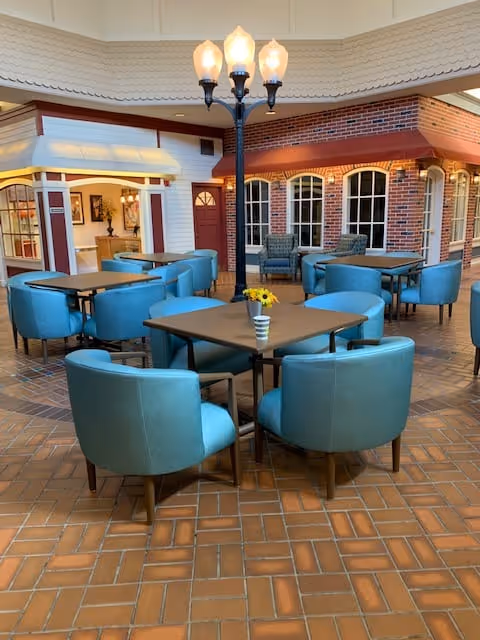 Indoor seating area with several square tables surrounded by teal upholstered chairs. The floor is tiled with a brick pattern. In the background, there is a decorative streetlamp with three lights and a facade resembling a small building with brick walls, windows, and a door. The ceiling is high and light-colored.