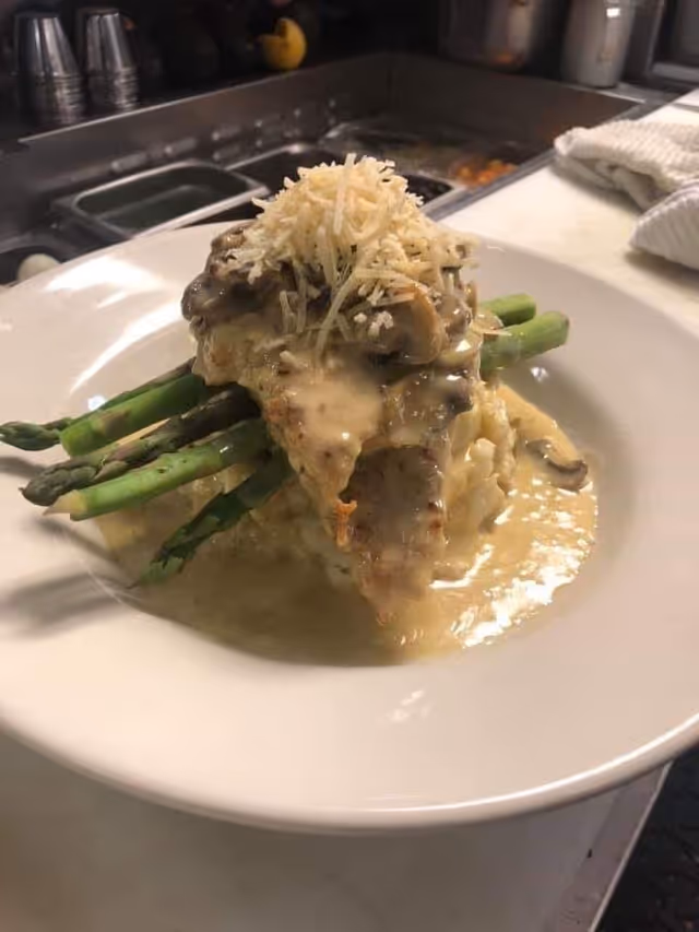 A plated dish featuring cooked asparagus spears topped with a creamy mushroom sauce, shredded cheese, and what appears to be a piece of chicken or fish, served on a white plate in a kitchen setting.