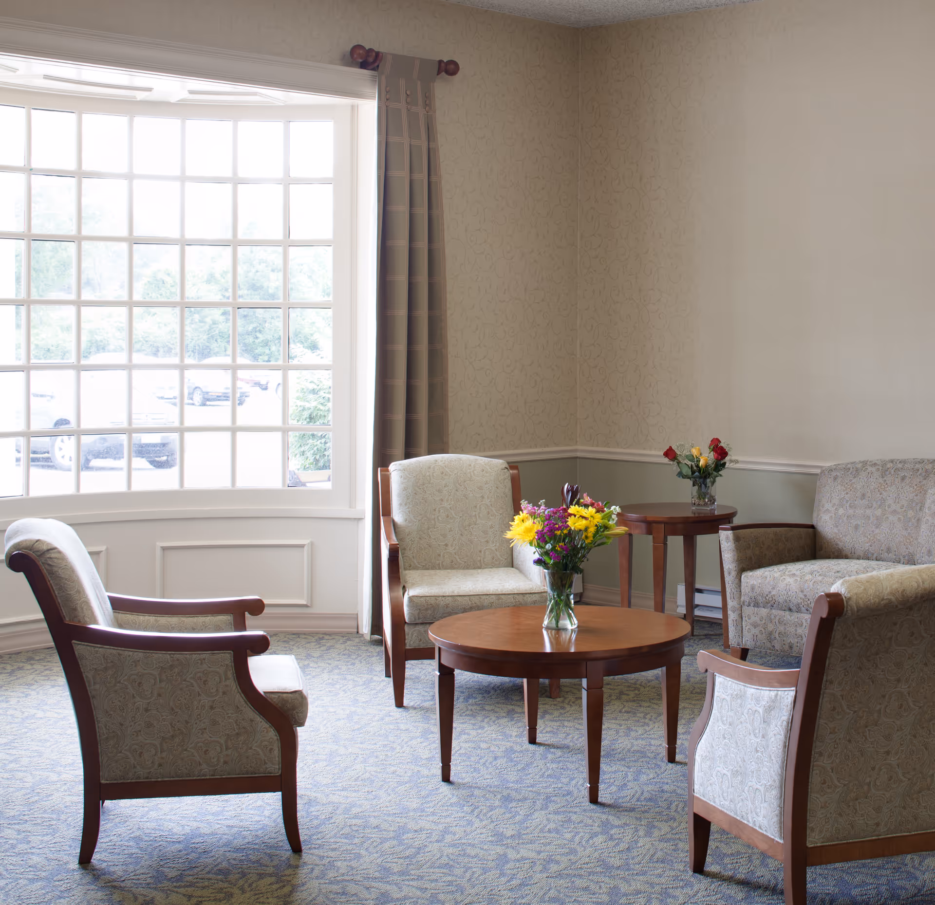 A cozy sitting area with four upholstered armchairs arranged around a round wooden coffee table with a vase of colorful flowers. There is a large window with multiple panes letting in natural light, and a small side table with another vase of flowers in the corner. The room has patterned carpet and wallpaper with a subtle design.