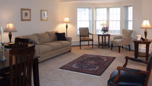 Bright living room with a beige sofa, several chairs around a bay window, floor lamps, and a patterned area rug.