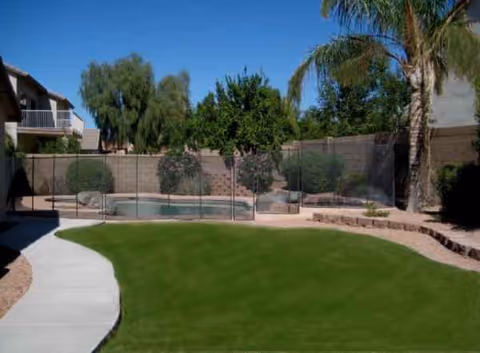 Sunny outdoor courtyard with a curved concrete path, green lawn, trees, and a fenced pool area.