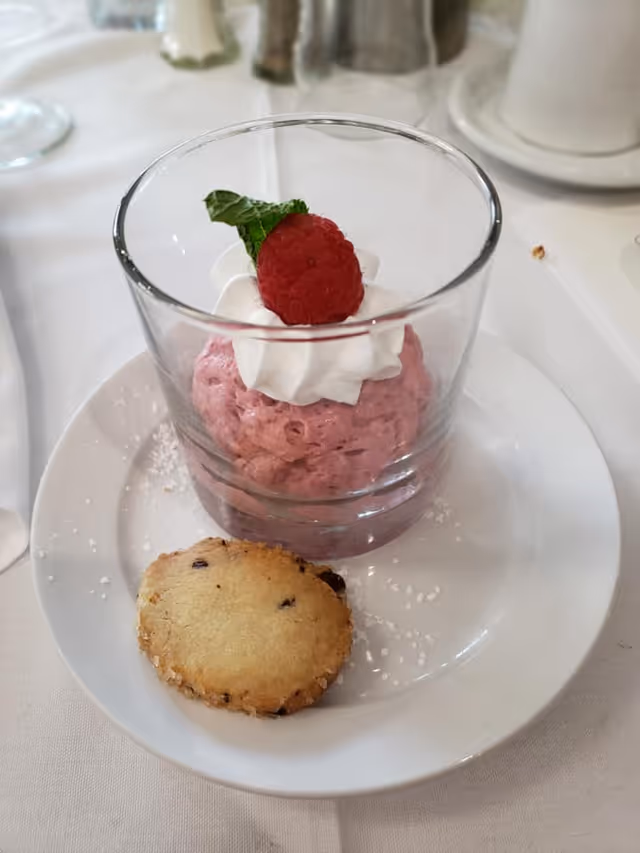 A dessert served on a white plate featuring a glass filled with pink mousse topped with whipped cream, a raspberry, and a mint leaf, accompanied by a cookie on the side. The plate is on a white tablecloth with some powdered sugar sprinkled around.