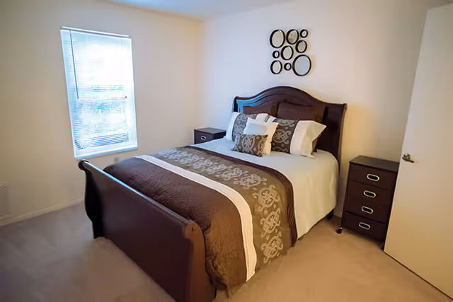 A bedroom with a wooden bed frame featuring a brown and beige patterned comforter and multiple pillows. There are two dark wooden nightstands on either side of the bed, one with drawers and the other with a single drawer. A window with blinds is on the left wall, and a decorative wall art piece with circular designs hangs above the bed.