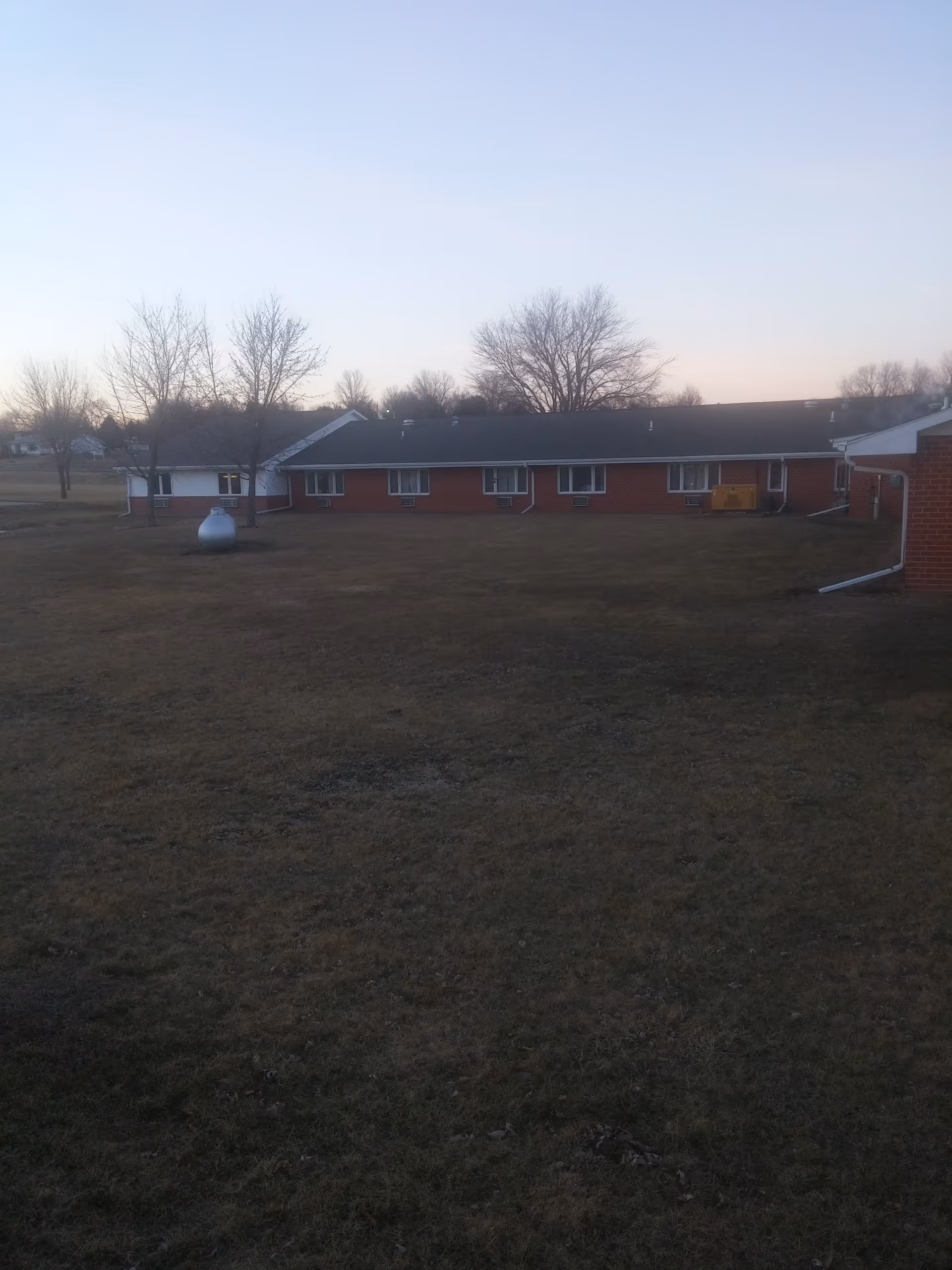 Single-story red-brick building viewed across a large grassy yard with bare trees under a pale evening sky.