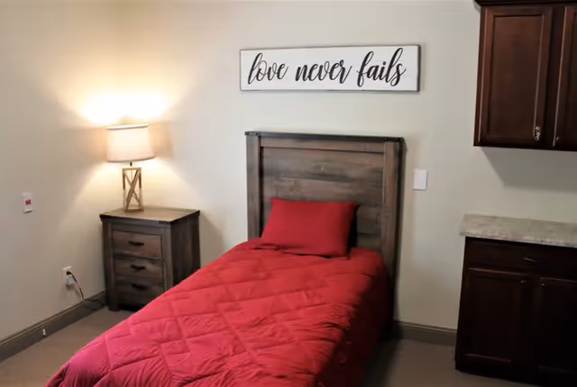 Single-occupancy bedroom with a twin bed covered in red bedding, wooden headboard, nightstand with a lamp, cabinets, and a wall sign that reads 'love never fails'.