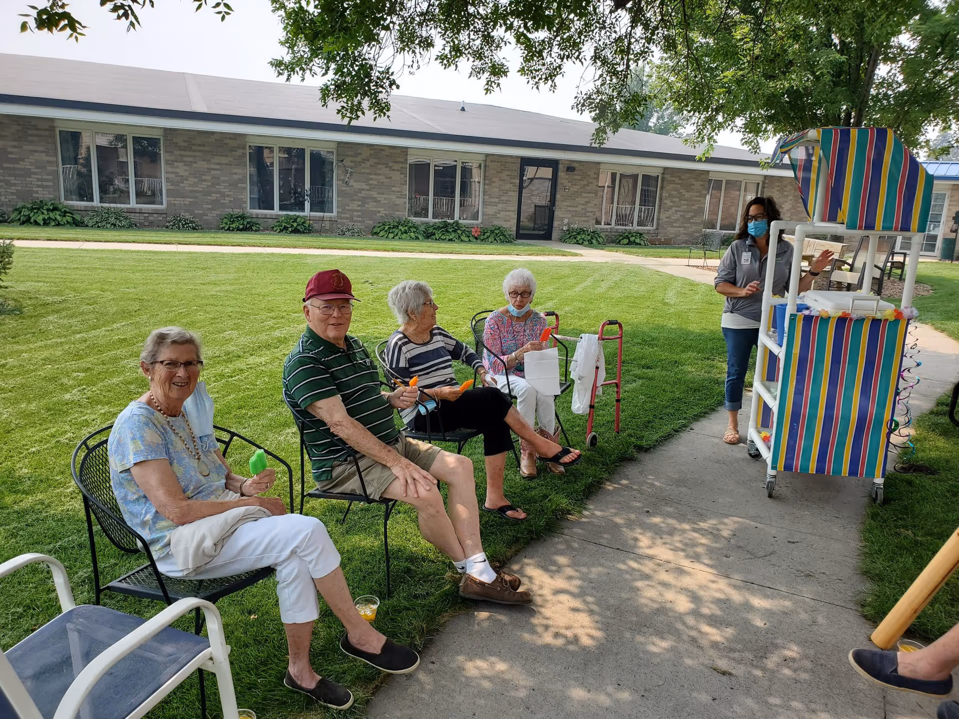 Four elderly people sitting on chairs outside on a lawn near a sidewalk, enjoying popsicles. A woman wearing a mask stands next to a colorful striped cart on the sidewalk. A single-story building with multiple windows is visible in the background.