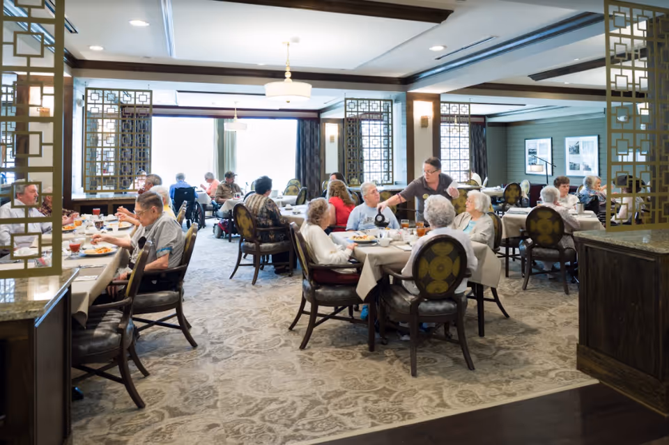 Seniors dining in a communal dining room with staff serving at round tables.