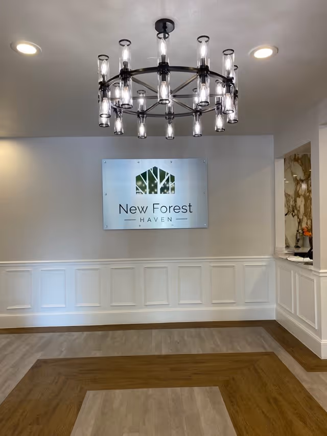Interior view of a room with a modern chandelier hanging from the ceiling, a sign on the wall reading 'New Forest Haven', white wainscoting on the walls, and a patterned wooden floor.