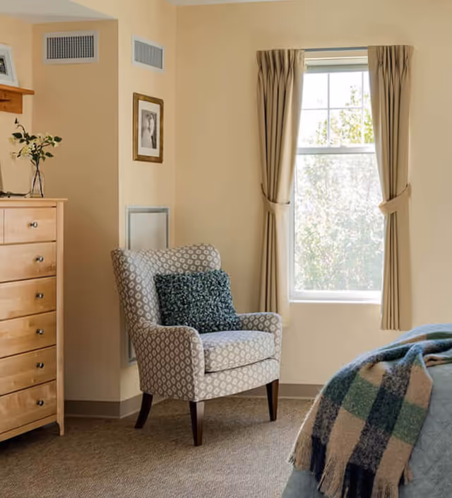 A cozy bedroom corner with a patterned armchair featuring a blue cushion, a wooden dresser with a vase of flowers on top, a window with beige curtains letting in natural light, and part of a bed with a blue and green plaid blanket.