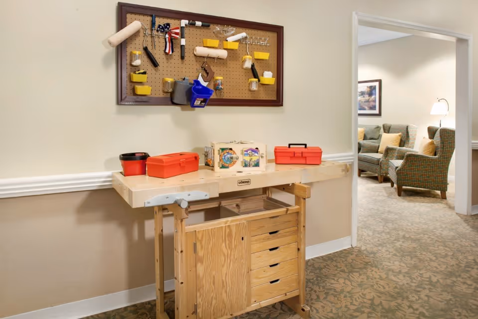 A wooden workbench with drawers and a pegboard above it holding various tools and small containers. On the workbench are two red toolboxes and a small activity box. In the background, there is a room with patterned armchairs, a floor lamp, and a framed picture on the wall.
