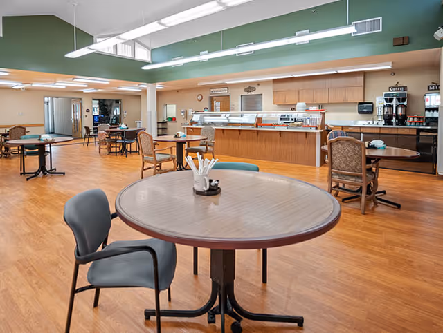 Spacious dining room with round tables and chairs facing a serving counter and coffee station.
