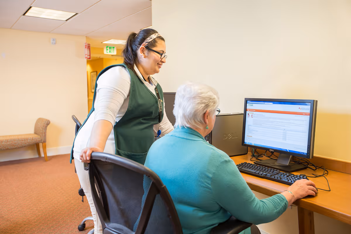 A staff member in a green vest assists an elderly woman seated at a desk using a desktop computer in a well-lit room with beige walls and carpeted floor.