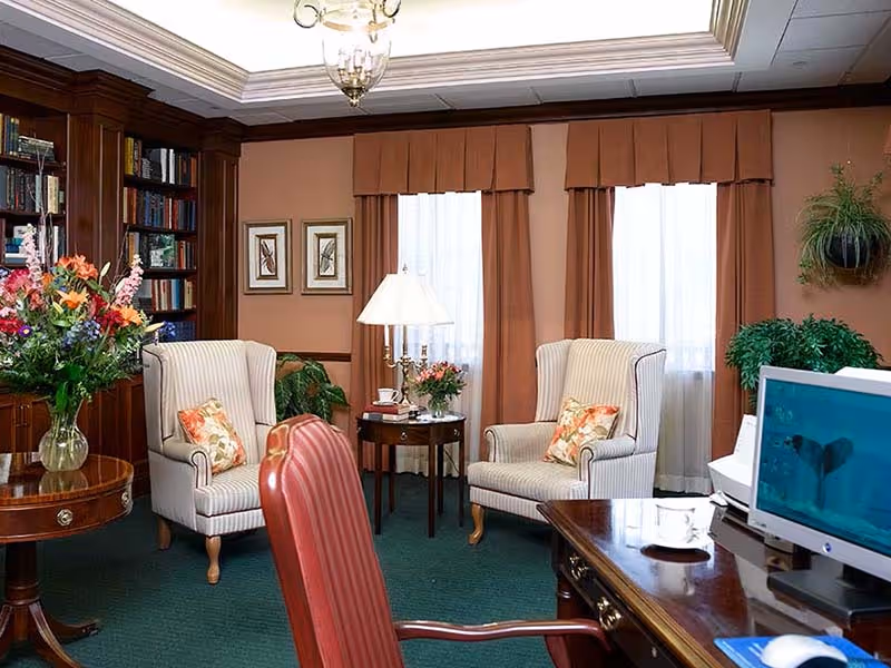 A cozy living room area with two striped armchairs each with a floral pillow, a wooden side table with a lamp and flowers, a wooden bookshelf filled with books, a round table with a vase of colorful flowers, and a desk with a computer monitor displaying an underwater scene. The room has green carpet, brown curtains, and framed bird artwork on the wall.