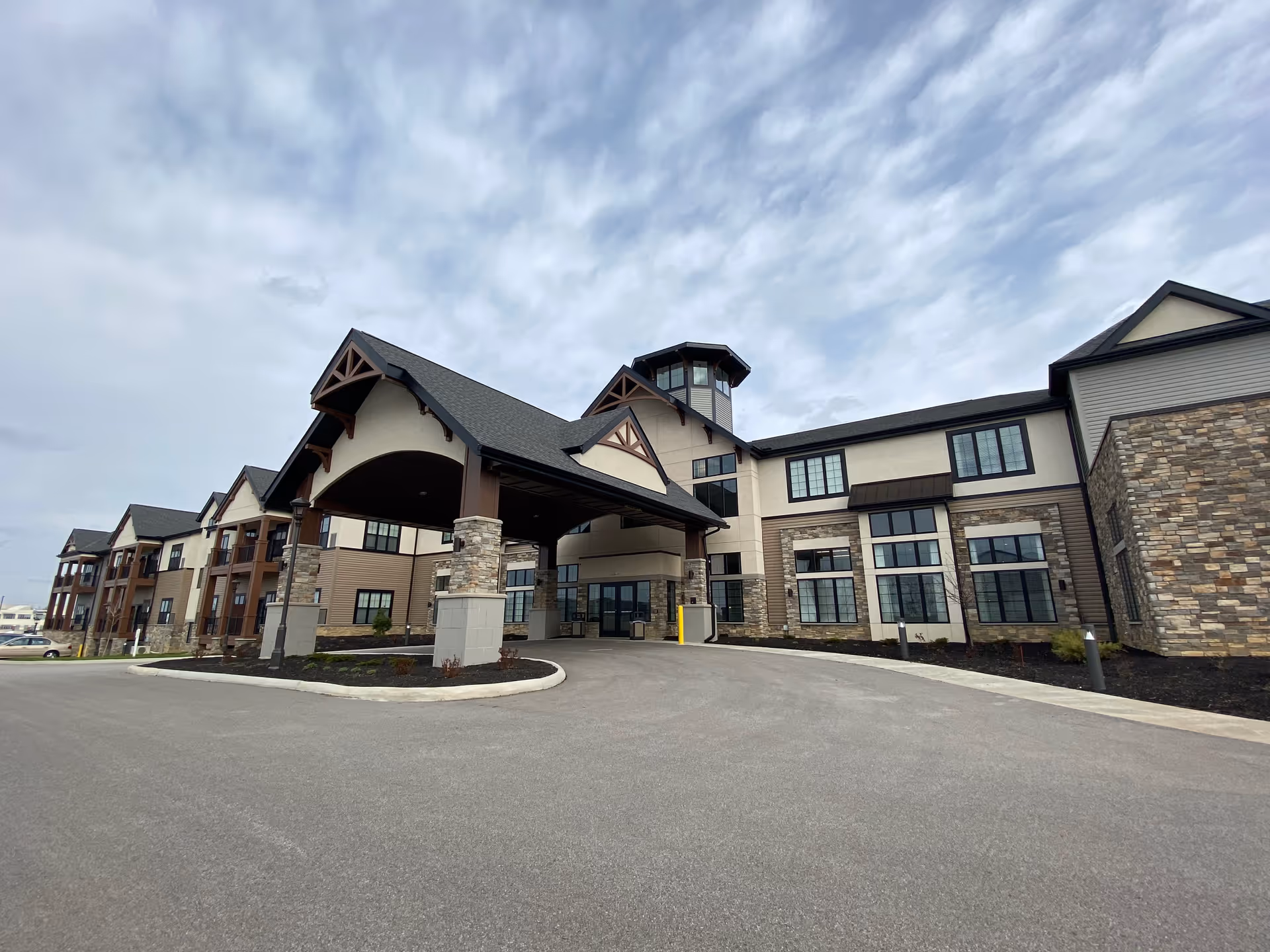 Exterior view of a large senior living facility building with a covered entrance supported by stone pillars, multiple windows, and a cloudy sky overhead.