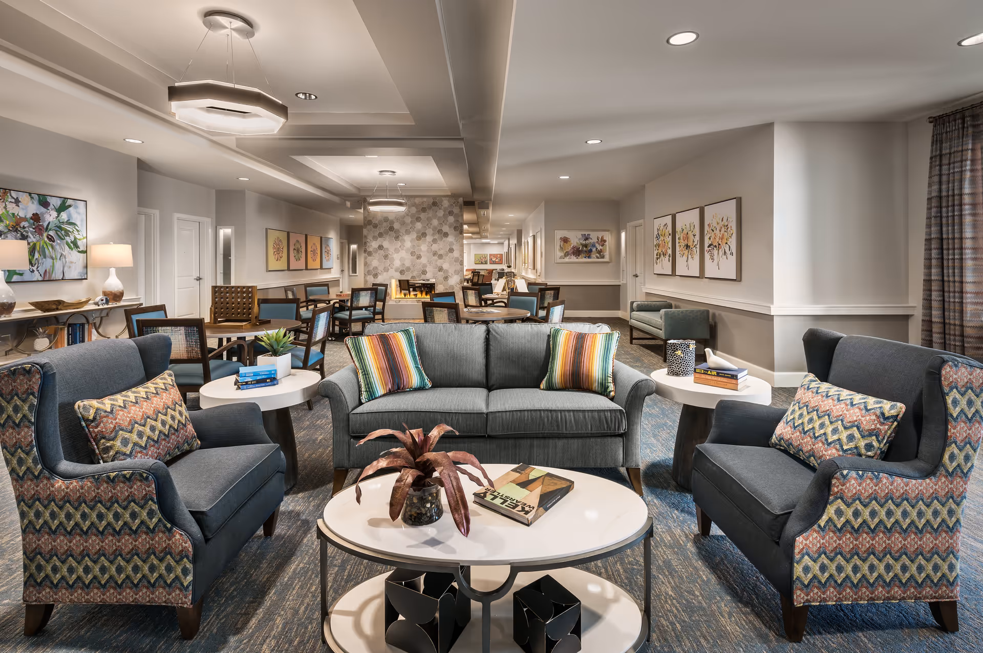 A spacious and well-lit assisted living common area with a gray sofa and two patterned armchairs arranged around a white coffee table with a plant and a book. The room features multiple round tables with chairs, colorful floral artwork on the walls, modern ceiling lights, and a cozy fireplace in the background.
