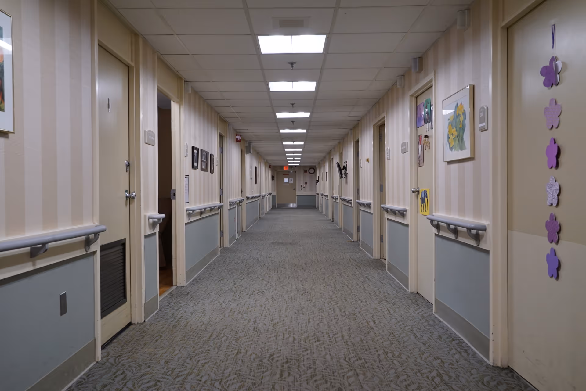 A long, well-lit hallway in a senior living facility with beige and gray walls, handrails on both sides, multiple closed doors, and some decorations and pictures on the walls.