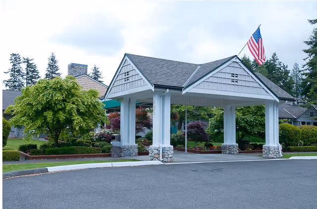 Covered entrance structure with white pillars and stone bases at a senior living facility, surrounded by well-maintained landscaping including trees and bushes, with an American flag flying on top.