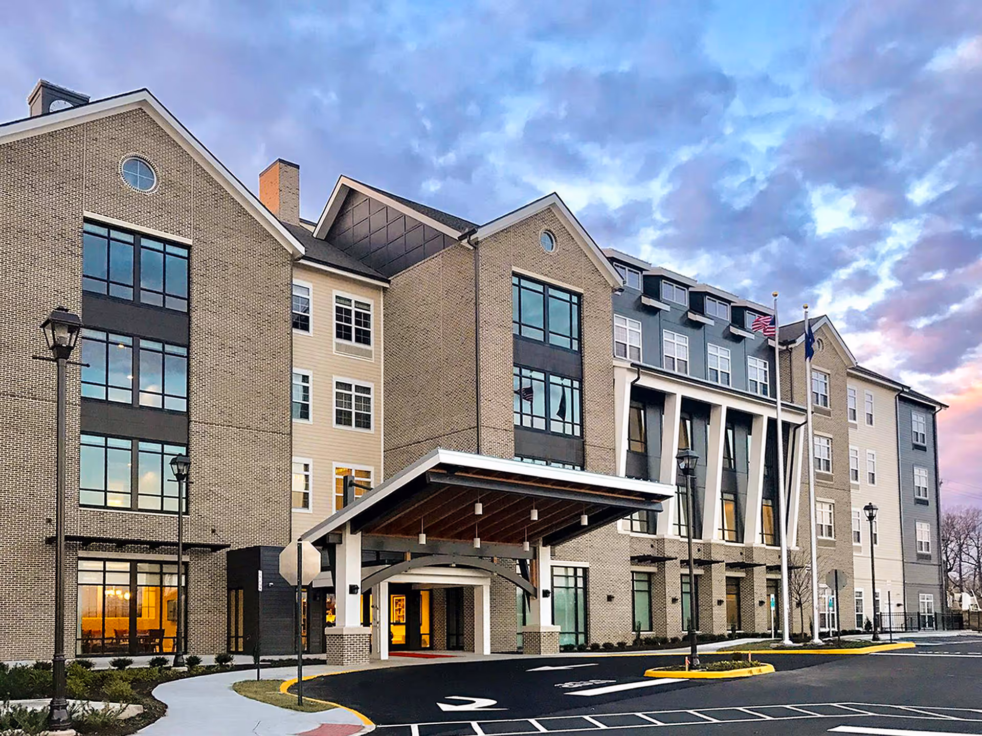 Exterior view of Tribute at The Glen senior living facility showing a multi-story building with large windows, a covered entrance, and two flagpoles with flags. The sky is partly cloudy during sunset.