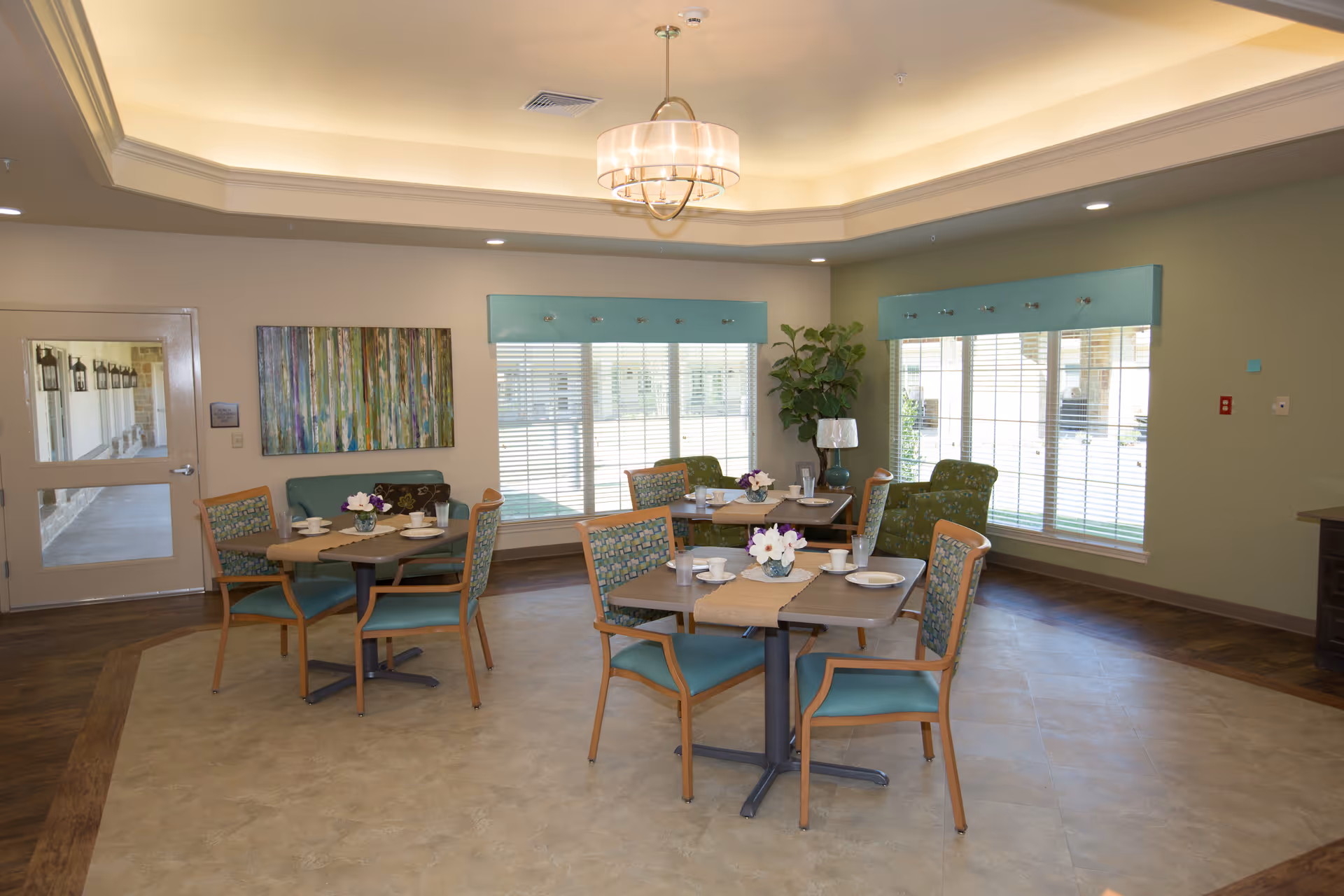 A dining room in a senior living facility with three tables, each set with plates, cups, and small flower arrangements. The room has large windows with teal valances, green walls, and a modern chandelier hanging from the ceiling. There are several chairs around the tables and a plant in the corner near the windows.
