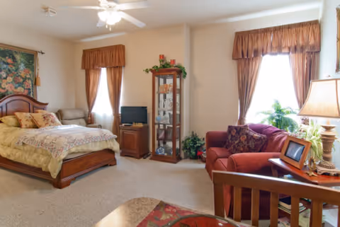 A cozy bedroom with a wooden bed featuring floral bedding, a small TV on a wooden stand, a glass display cabinet with decorative items, a red upholstered armchair with patterned cushions, a wooden side table with a lamp and picture frame, and two windows with brown curtains allowing natural light to enter.
