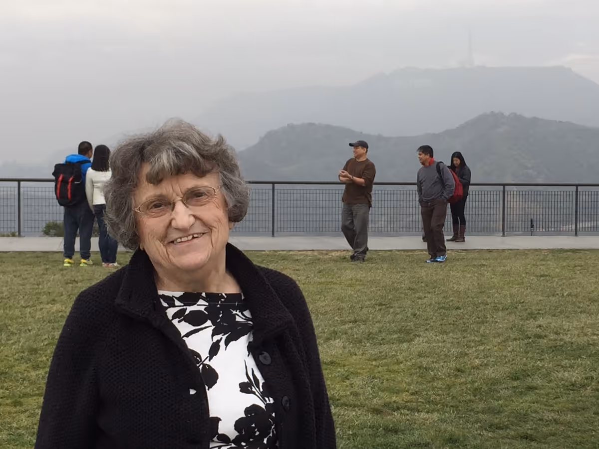 An elderly woman with glasses and curly gray hair smiling at the camera, standing on a grassy area with a railing and mountains in the background. Several people are also visible in the background near the railing.