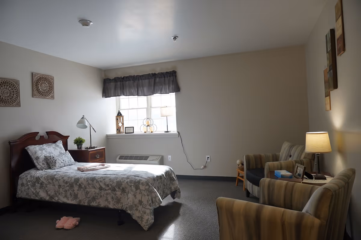 A cozy bedroom in a senior living facility with a single bed covered in a floral patterned bedspread. Next to the bed is a wooden nightstand with a lamp and a small plant. A window with a dark valance lets in natural light. Two decorative wall hangings are above the bed. Across from the bed are two striped armchairs with a small table and lamp between them. The room has neutral-colored walls and carpeted floor.