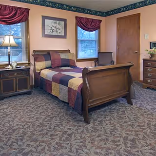 Cozy single bed in a decorated senior living bedroom with wooden furniture, patterned quilt, windows with valances, and carpeted floor.