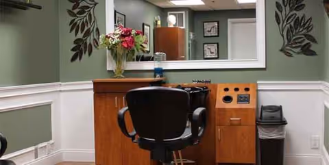 A salon styling station with a black salon chair in front of a large mirror and wooden cabinet, decorated with flowers and wall decals.