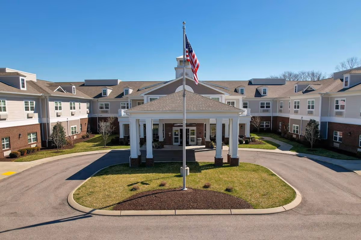 Front entrance of a senior living building with a covered porte-cochère, circular driveway, and an American flag flying in front.