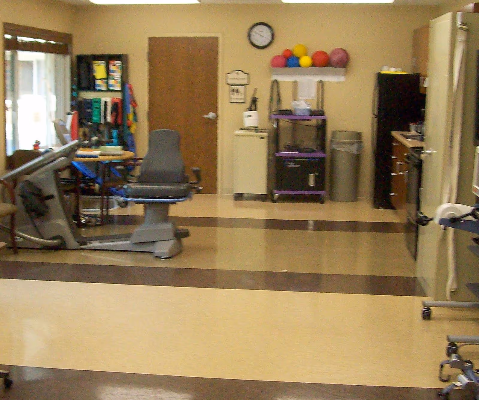 Interior room with exercise equipment including a recumbent bike, therapy balls on a shelf, resistance bands, and other physical therapy tools. The room has a tiled floor with brown and beige stripes, a clock on the wall, and a closed wooden door in the background.