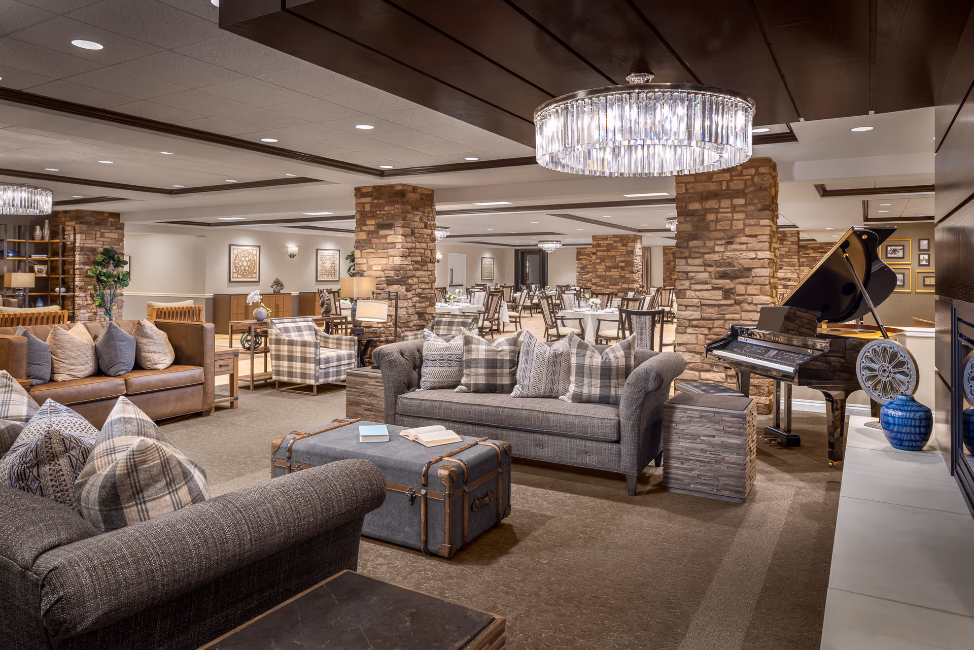 A spacious senior living common area featuring comfortable sofas with patterned pillows, a vintage-style trunk coffee table, a grand piano, and stone pillars. In the background, there is a dining area with multiple tables and chairs. The room is warmly lit with modern chandeliers and recessed ceiling lights.