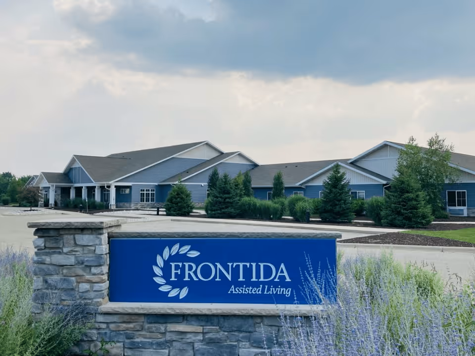Exterior view of Frontida Assisted Living facility showing a large building with blue siding and a stone sign in the foreground with the facility name. The area is landscaped with bushes, trees, and purple flowering plants under a cloudy sky.