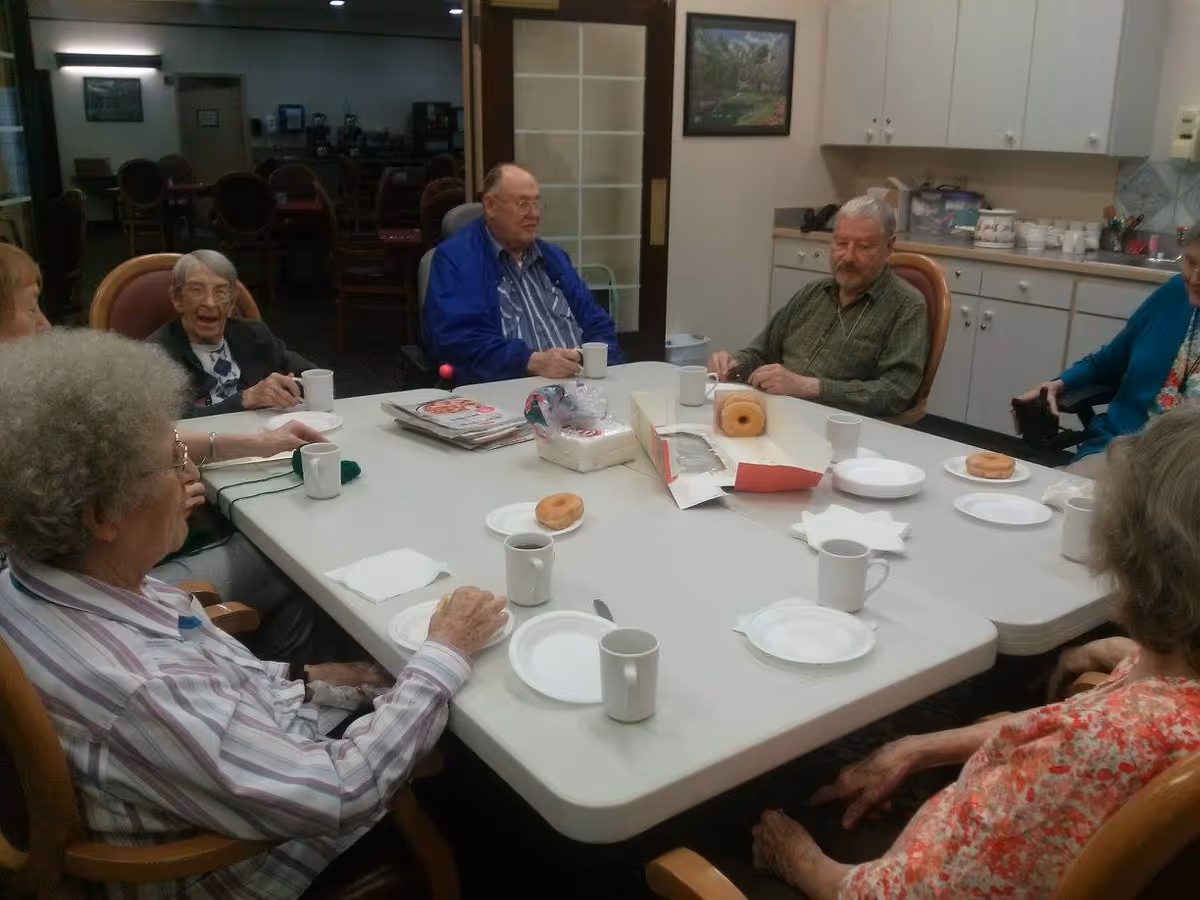A group of elderly people sitting around a rectangular table in a communal room, enjoying coffee and donuts. The table has white plates, coffee mugs, a box of donuts, and some napkins. The background shows a kitchenette area with cabinets and a counter.