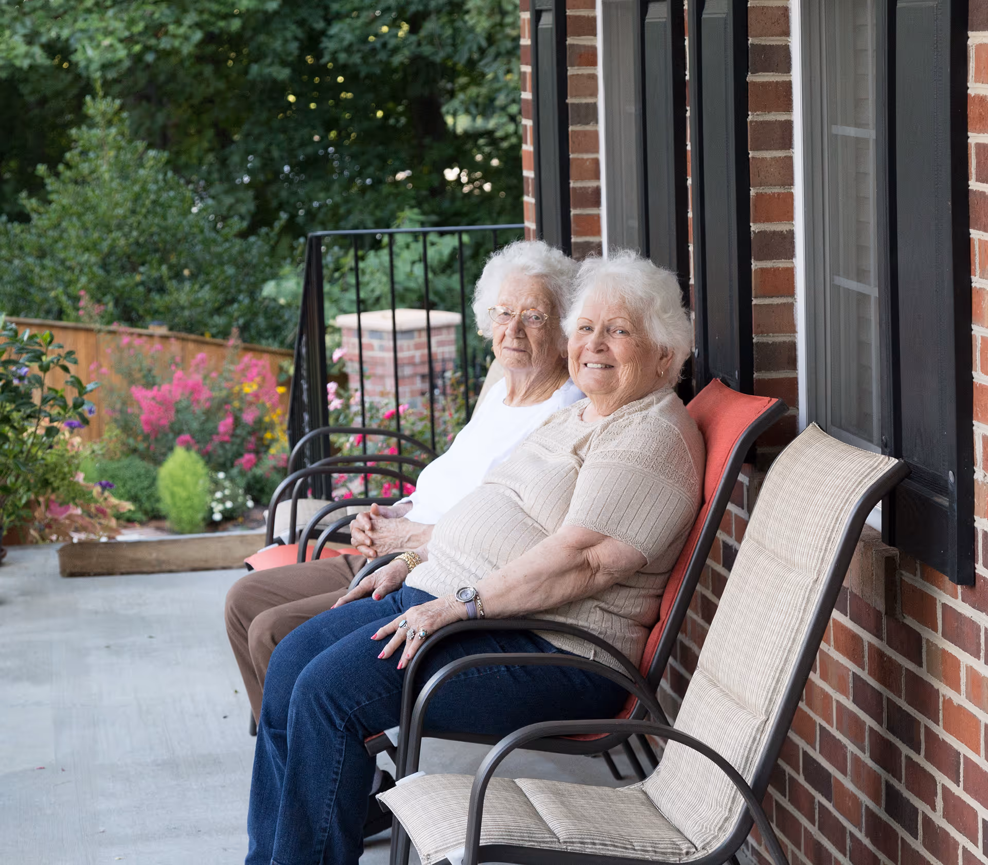 Two elderly women sitting on chairs on a porch outside a brick building, with greenery and colorful flowers in the background.